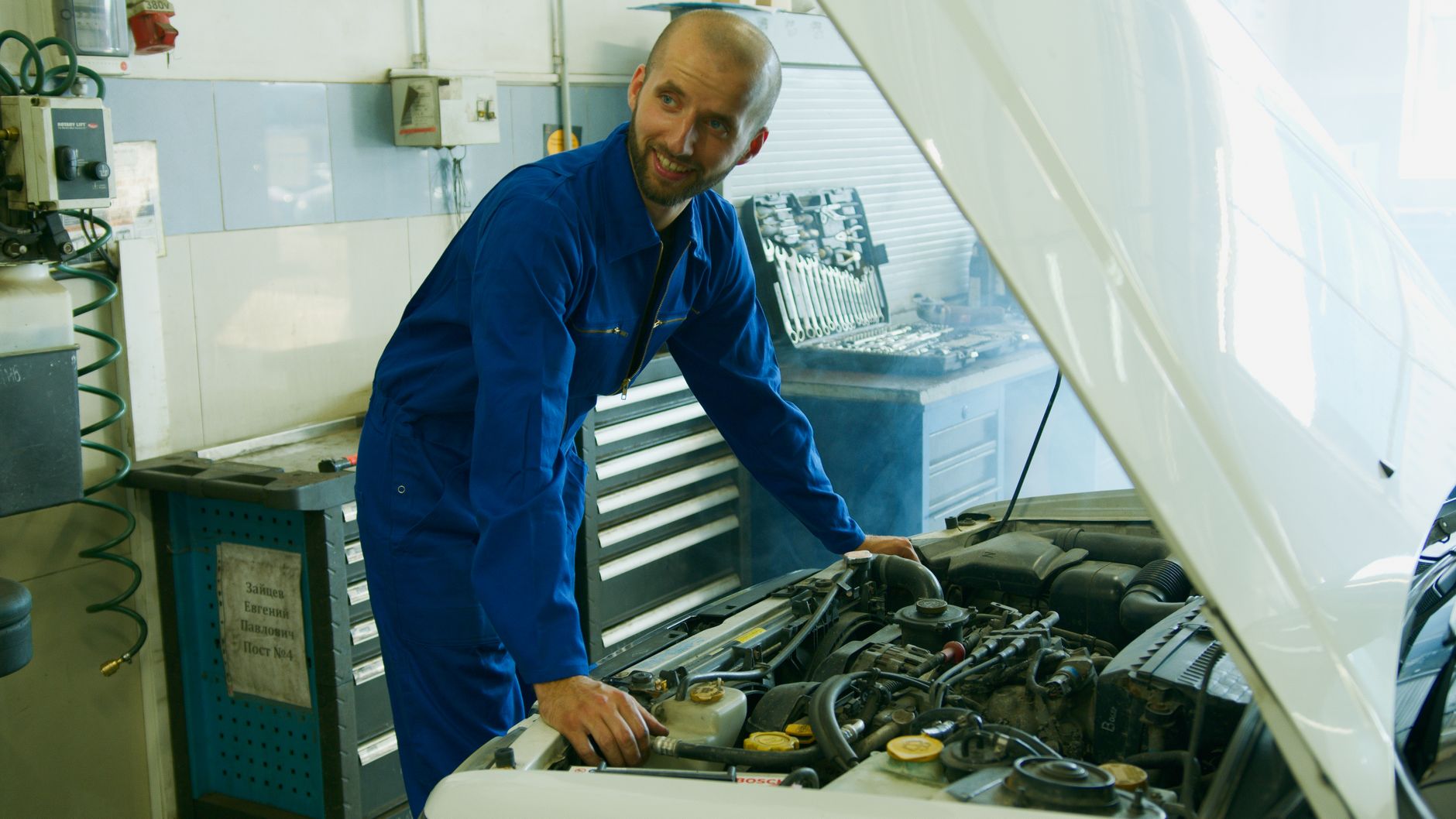 Smiling mechanic in a blue overall inspecting a car engine