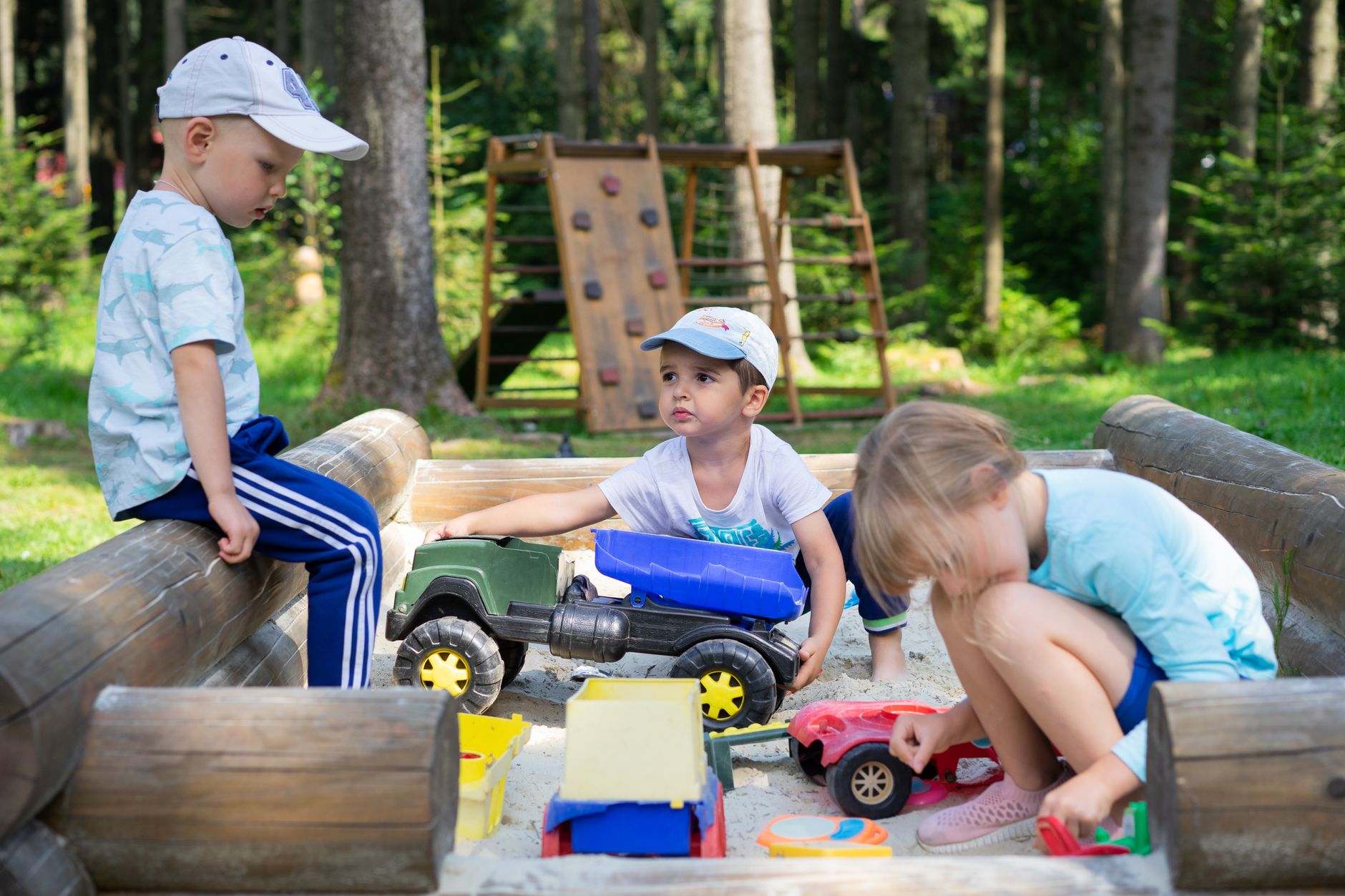 Three children engaged in imaginative play in an outdoor sandbox with toys.