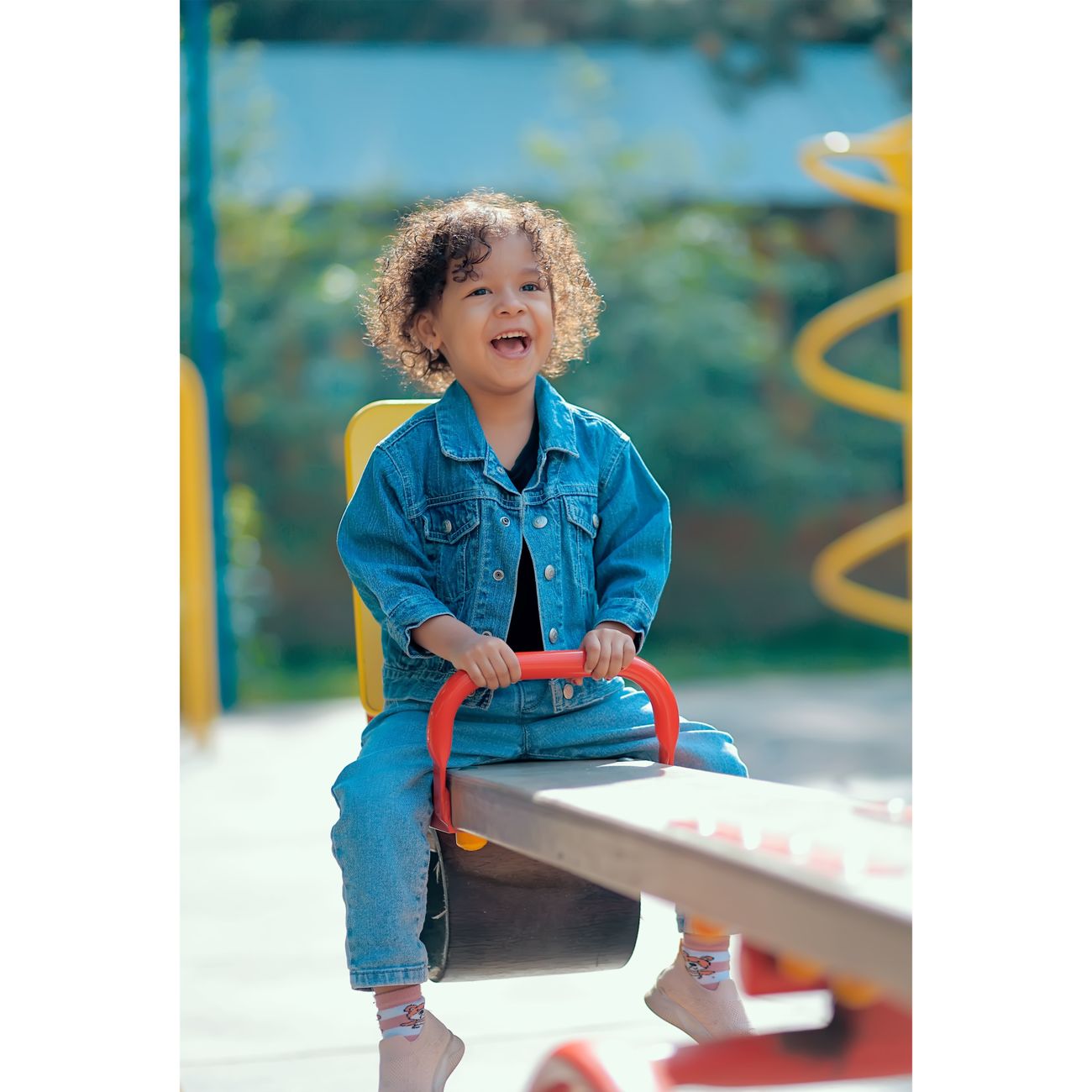 A happy child with curly hair enjoys a fun moment on a playground seesaw wearing a denim jacket.