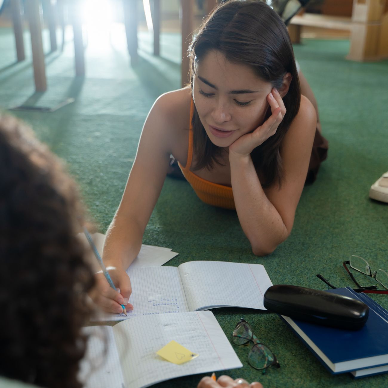 Casual study session: young woman writing notes with a classmate indoors.